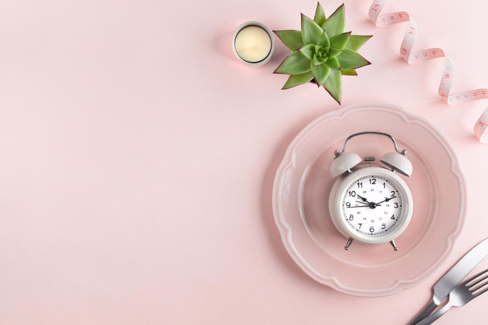 A flat lay of a pink plate with a vintage alarm clock on it, next to a succulent plant and a measuring tape, symbolizing meal timing or fasting.