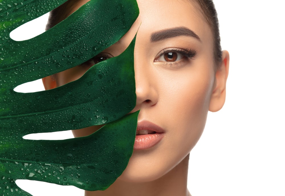 A close-up of a woman partially covered by a large green leaf, highlighting chlorella's moisturizing and detoxifying effects on the skin.