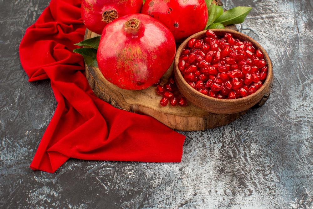 Whole pomegranates and a bowl of seeds, sources of ellagic acid, on a red cloth.