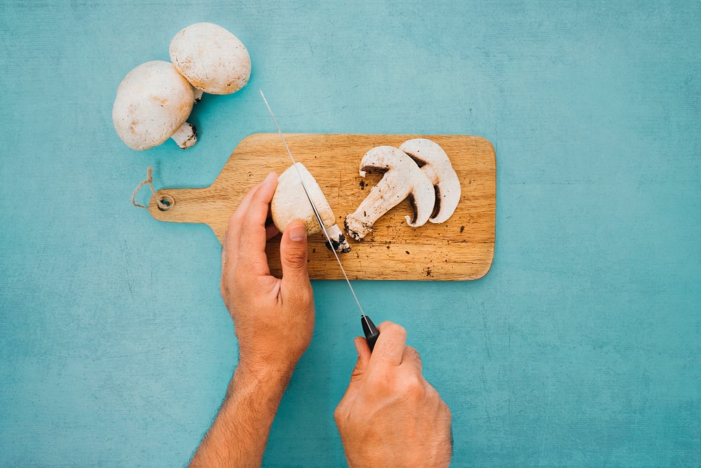 A person slicing white button mushrooms on a wooden cutting board with a knife.